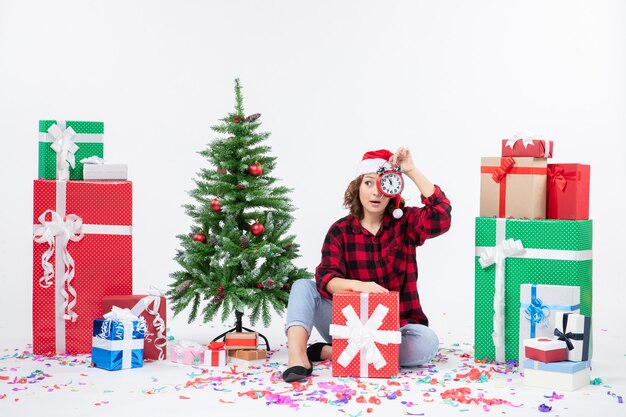 Vista frontal de la mujer joven sentada alrededor de regalos de Navidad sosteniendo relojes en la pared blanca
