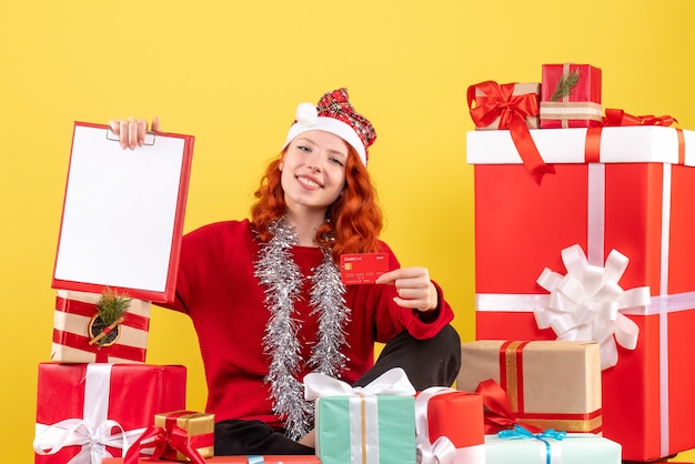 Vista frontal de la mujer joven sentada alrededor de regalos de Navidad con tarjeta bancaria en la pared amarilla