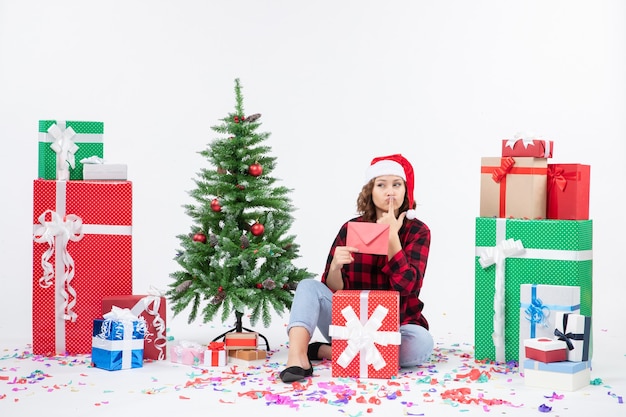 Vista frontal de la mujer joven sentada alrededor de regalos sosteniendo envolver en la pared blanca