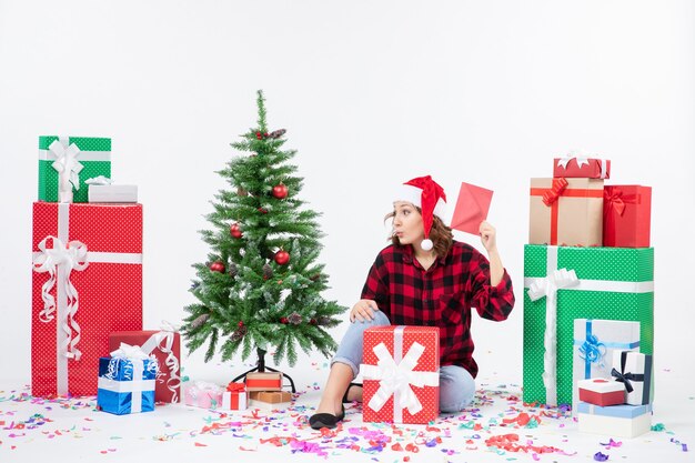 Vista frontal de la mujer joven sentada alrededor de regalos sosteniendo envolver en la pared blanca