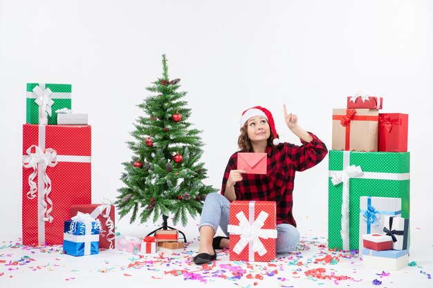 Vista frontal de la mujer joven sentada alrededor de regalos sosteniendo envolver en la pared blanca