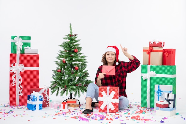 Vista frontal de la mujer joven sentada alrededor de regalos sosteniendo envolver en la pared blanca