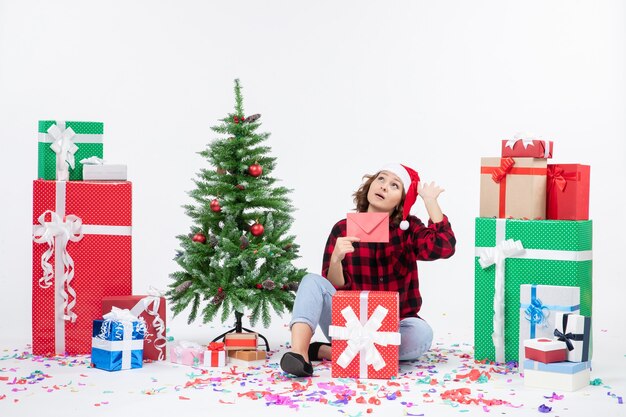 Vista frontal de la mujer joven sentada alrededor de regalos sosteniendo envolver en la pared blanca