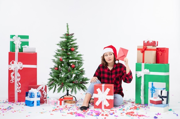 Vista frontal de la mujer joven sentada alrededor de regalos sosteniendo envolver en la pared blanca