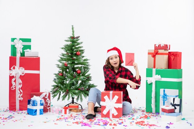 Vista frontal de la mujer joven sentada alrededor de regalos sosteniendo envolver en la pared blanca