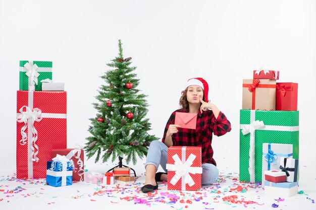 Vista frontal de la mujer joven sentada alrededor de regalos sosteniendo envolver en la pared blanca