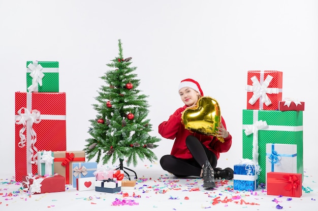 Vista frontal de la mujer joven sentada alrededor de regalos sosteniendo la figura del corazón de oro en la pared blanca