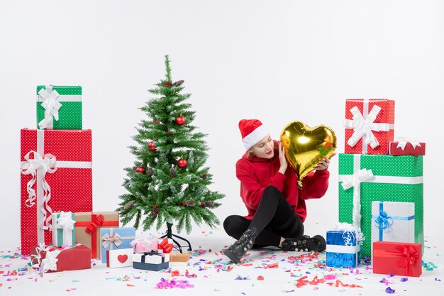 Vista frontal de la mujer joven sentada alrededor de regalos sosteniendo la figura del corazón de oro en la pared blanca