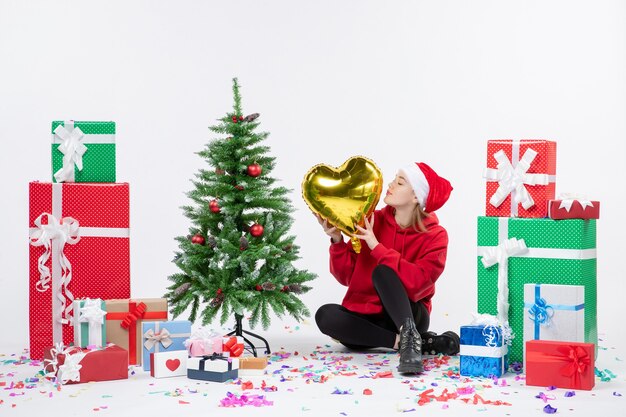 Vista frontal de la mujer joven sentada alrededor de regalos sosteniendo la figura del corazón de oro en la pared blanca