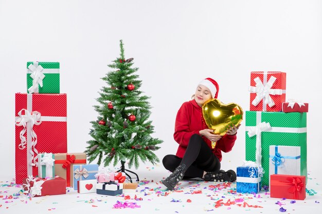 Vista frontal de la mujer joven sentada alrededor de regalos sosteniendo la figura del corazón de oro en la pared blanca