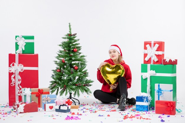 Vista frontal de la mujer joven sentada alrededor de regalos sosteniendo la figura del corazón de oro en la pared blanca