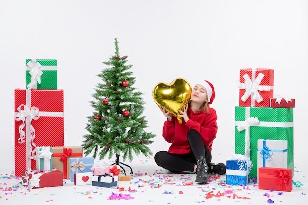 Vista frontal de la mujer joven sentada alrededor de regalos sosteniendo una figura en forma de corazón de oro en la pared blanca