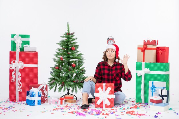 Vista frontal de la mujer joven sentada alrededor de regalos sosteniendo relojes en la cabeza en la pared blanca