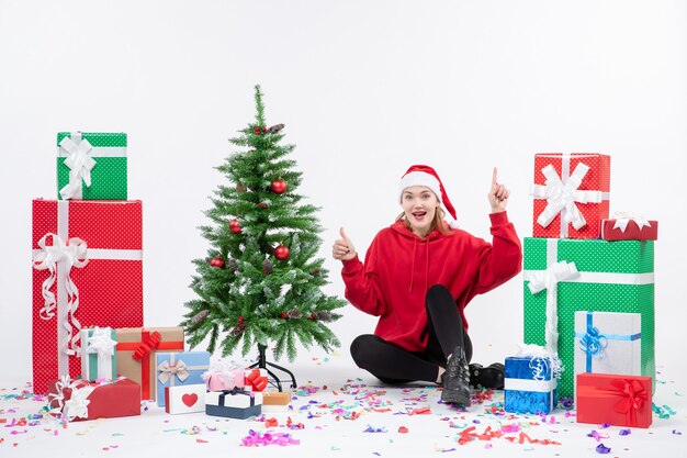 Vista frontal de la mujer joven sentada alrededor de regalos de vacaciones emocionados en la pared blanca