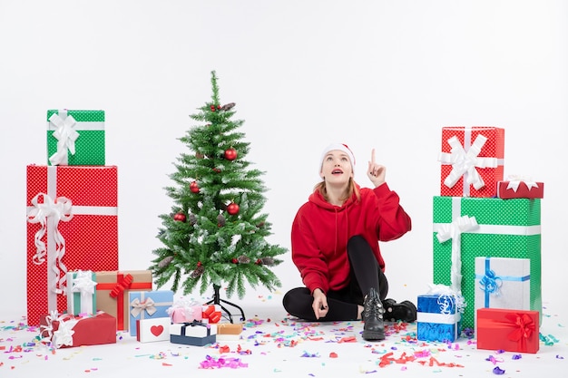 Vista frontal de la mujer joven sentada alrededor de regalos de vacaciones en la pared blanca