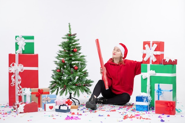 Vista frontal de la mujer joven sentada alrededor de regalos de vacaciones en la pared blanca