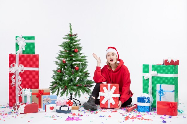 Vista frontal de la mujer joven sentada alrededor de regalos de vacaciones en la pared blanca