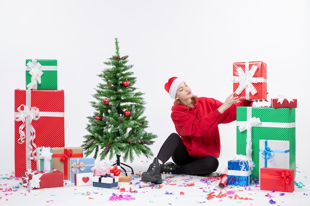 Vista frontal de la mujer joven sentada alrededor de regalos de vacaciones en la pared blanca