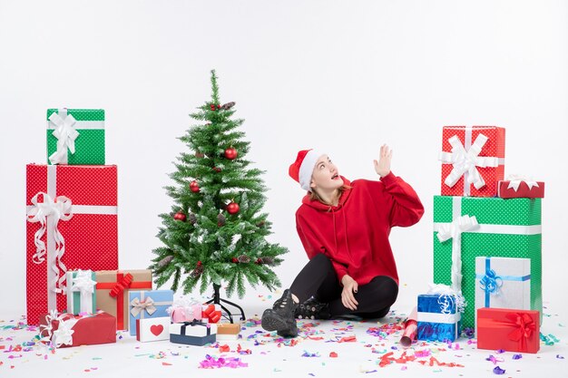 Vista frontal de la mujer joven sentada alrededor de regalos de vacaciones en la pared blanca