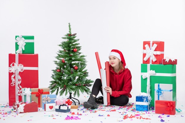 Vista frontal de la mujer joven sentada alrededor de regalos de vacaciones en la pared blanca