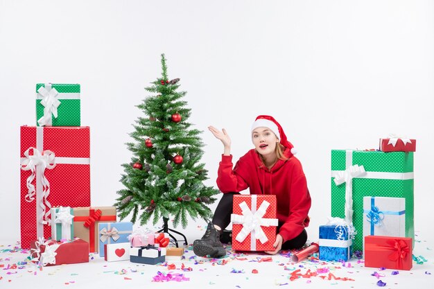 Vista frontal de la mujer joven sentada alrededor de regalos de vacaciones en la pared blanca