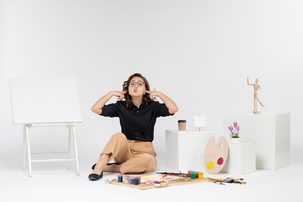 Vista frontal mujer joven sentada dentro de la habitación con caballete y pinturas sobre fondo blanco.