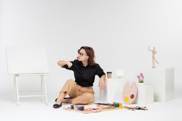 Vista frontal mujer joven sentada dentro de la habitación con caballete y pinturas sobre fondo blanco.