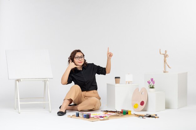 Vista frontal mujer joven sentada dentro de la habitación con caballete y pinturas sobre fondo blanco.