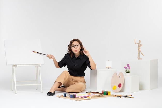 Vista frontal mujer joven sentada dentro de la habitación con pinturas y caballete sobre fondo blanco.