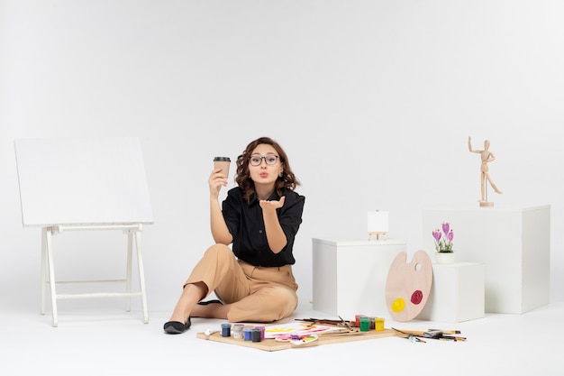 Vista frontal mujer joven sentada dentro de la habitación con pinturas y caballete sobre fondo blanco.