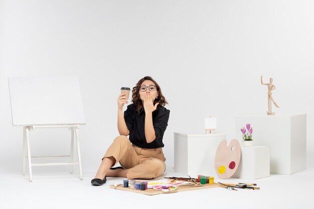 Vista frontal mujer joven sentada dentro de la habitación con pinturas y caballete sobre fondo blanco.