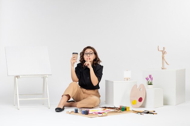 Vista frontal mujer joven sentada dentro de la habitación con pinturas y caballete sobre fondo blanco.