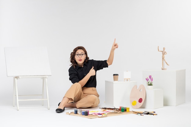 Vista frontal mujer joven sentada dentro de la habitación con pinturas y caballete sobre fondo blanco.