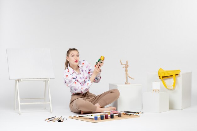 Vista frontal mujer joven sentada dentro de la habitación con pinturas y caballete sobre fondo blanco.