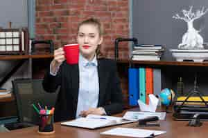 Foto gratuita vista frontal de la mujer joven sentada en una mesa y sosteniendo una taza roja posando para la cámara en la oficina