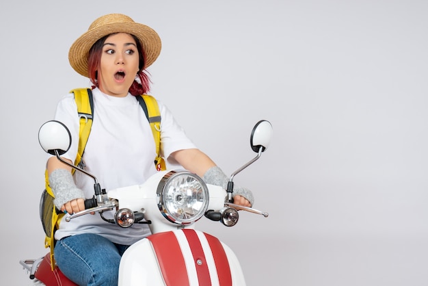 Vista frontal mujer joven sentada en motocicleta con mochila y sombrero pared blanca