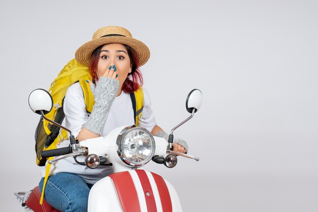 Vista frontal mujer joven sentada en motocicleta con mochila y sombrero pared blanca