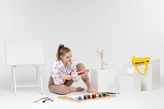 Vista frontal mujer joven sentada con pinturas y caballete para dibujar en el escritorio blanco dibujar mujer pintura artista pintor color de arte