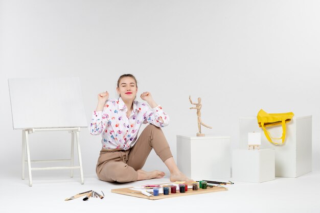 Vista frontal mujer joven sentada con pinturas y caballete para dibujar sobre fondo blanco.