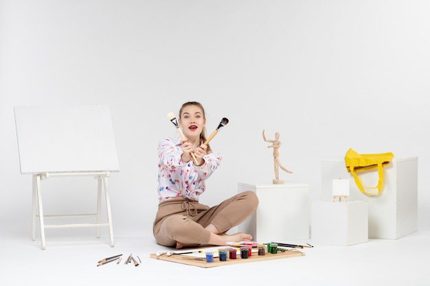 Vista frontal mujer joven sentada con pinturas y caballete para dibujar sobre fondo blanco.