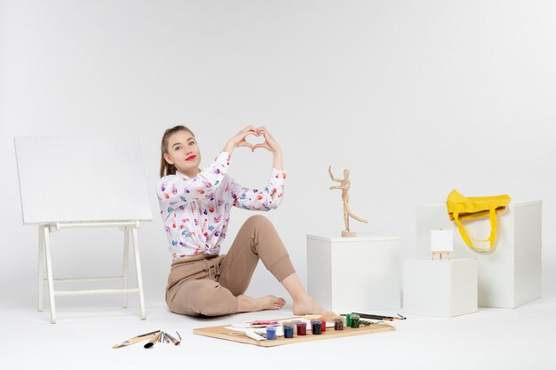 Vista frontal mujer joven sentada con pinturas y caballete para dibujar sobre fondo blanco.