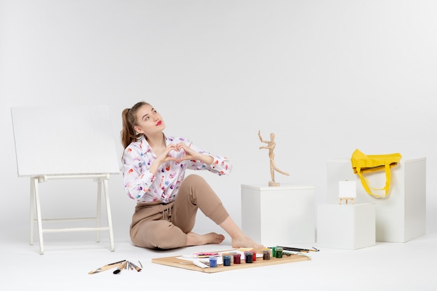 Vista frontal mujer joven sentada con pinturas y caballete para dibujar sobre fondo blanco.