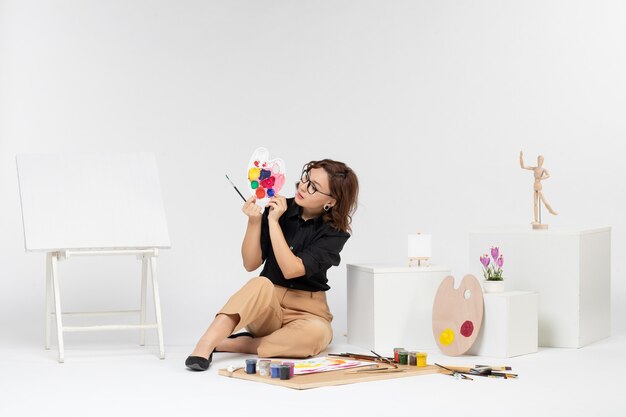 Vista frontal mujer joven sentada con pinturas y caballete sobre fondo blanco.