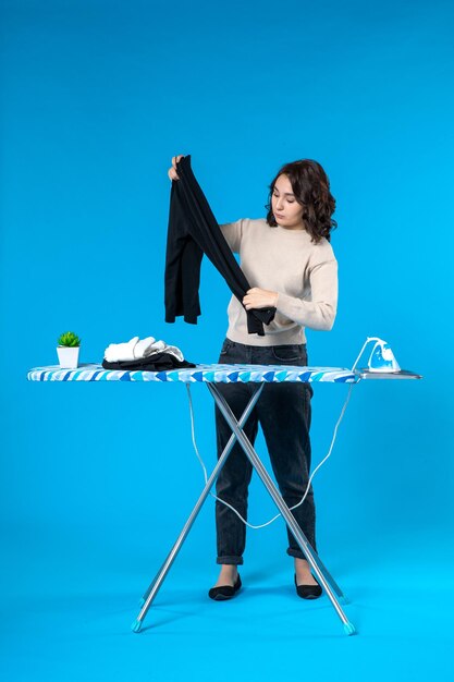 Vista frontal de una mujer joven seria de pie detrás de la tabla de planchar y sosteniendo un paño sobre la superficie de la onda azul