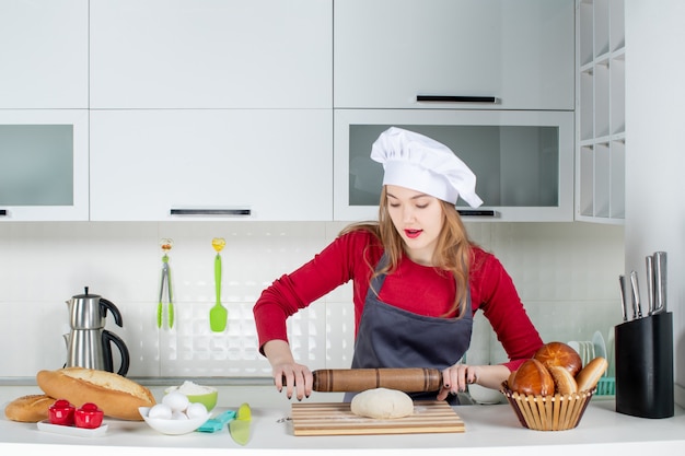 Vista frontal de la mujer joven con sombrero de cocinero y delantal de laminación de masa en la cocina