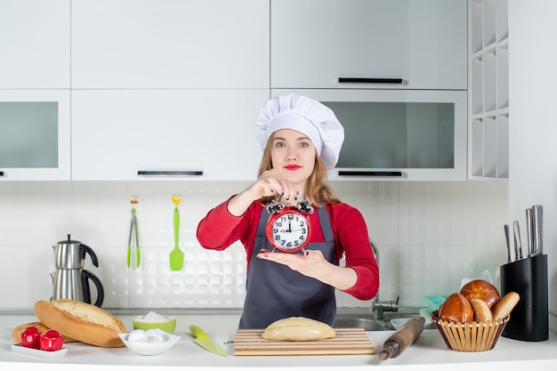 Vista frontal de la mujer joven con sombrero de cocinero y delantal con reloj despertador rojo en la cocina