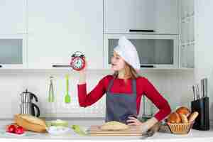 Foto gratuita vista frontal de la mujer joven con sombrero de cocinero y delantal con reloj despertador rojo poniendo la mano en la cintura en la cocina