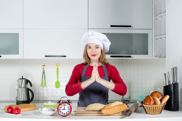 Vista frontal de la mujer joven con sombrero de cocinero y delantal uniendo las manos en la cocina