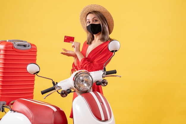 Vista frontal de la mujer joven con sombrero de Panamá con tarjeta de crédito cerca de ciclomotor