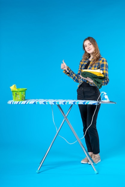 Foto gratuita vista frontal de la mujer joven sonriente sosteniendo la ropa doblada haciendo un gesto de ok y de pie detrás de la tabla de planchar en el lavadero